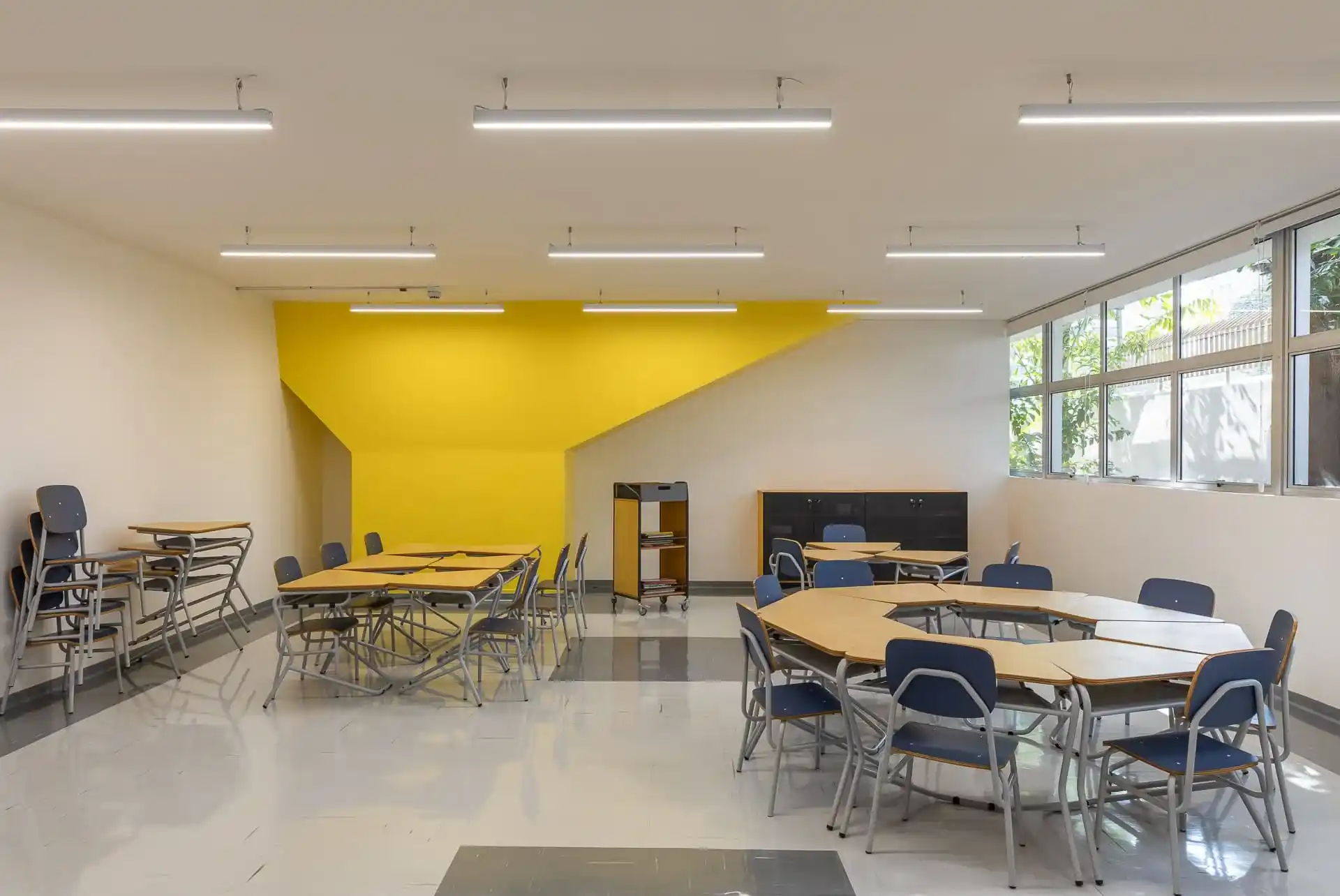 Sala de aula com carteiras escolares com tampo em formato de trapézio, distribuídas pelo ambiente com cadeiras formando conjuntos variados.