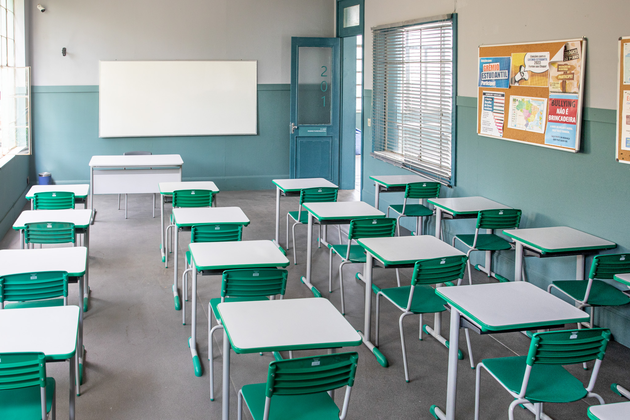 Sala de aula com mesas e cadeiras escolares em cor verde, ao fundo parede pintada até a metade da altura de verde com lousa branca