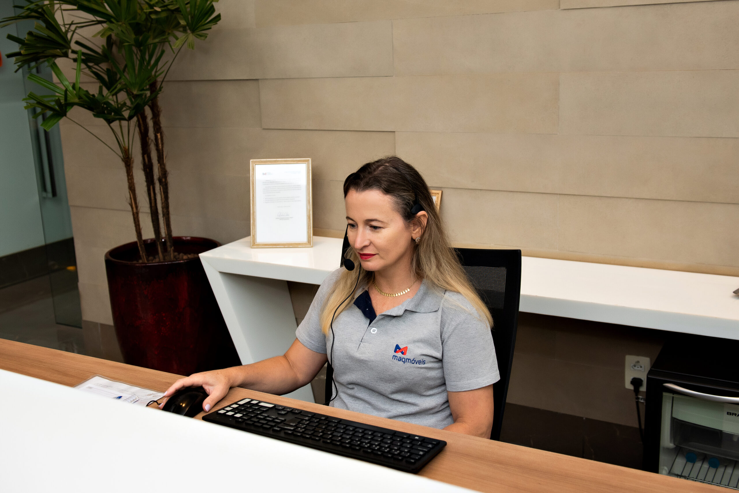 A imagem apresenta uma mulher de pele clara e cabelos loiros, vestindo uma camiseta cinza em seu posto de trabalho, atuando como recepcionista