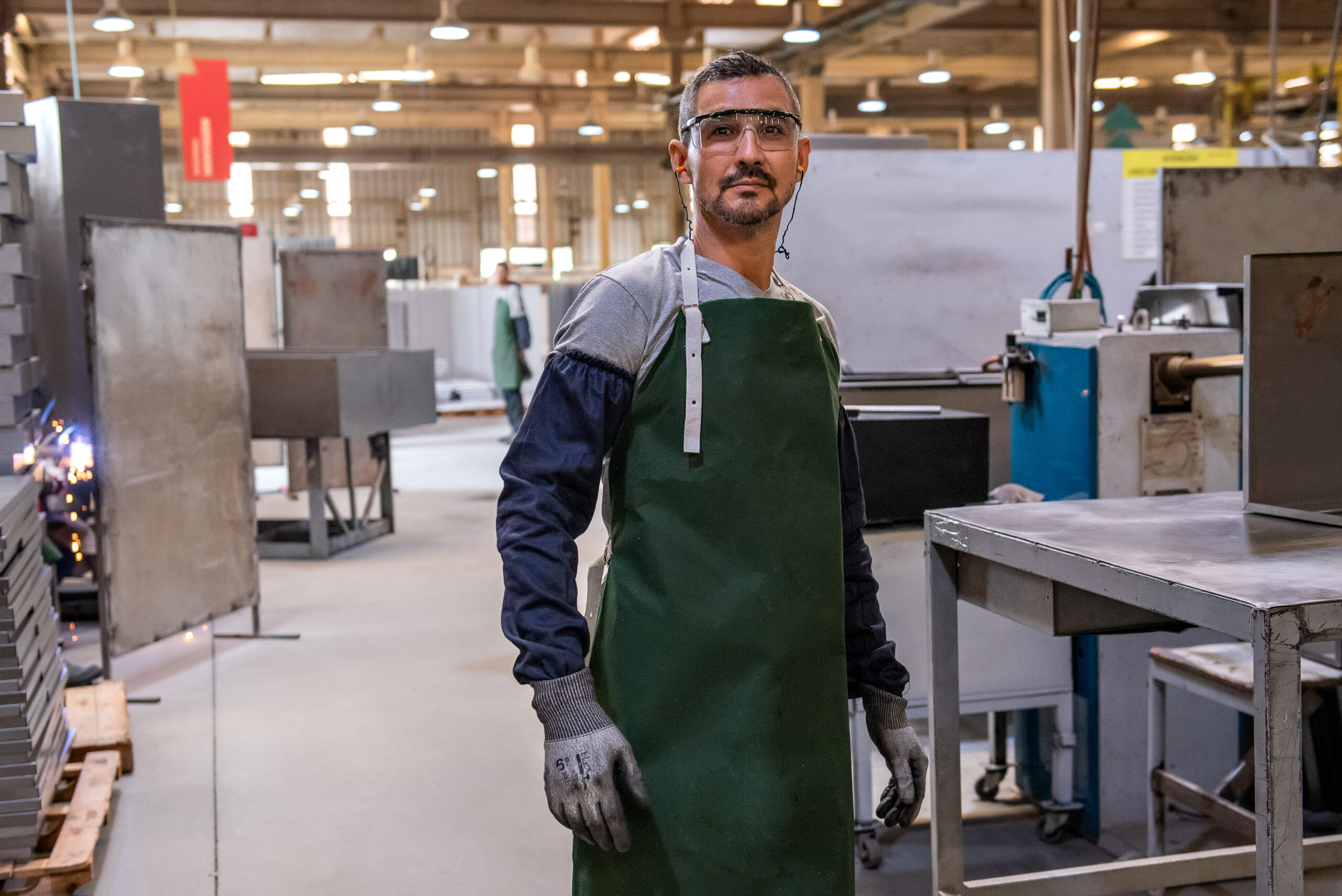 A imagem apresenta em primeiro plano um homem de pele clara e cabelos grisalhos, vestido avental, luvas e viseiras de segurança, enquanto exerce seu trabalho em uma fábrica