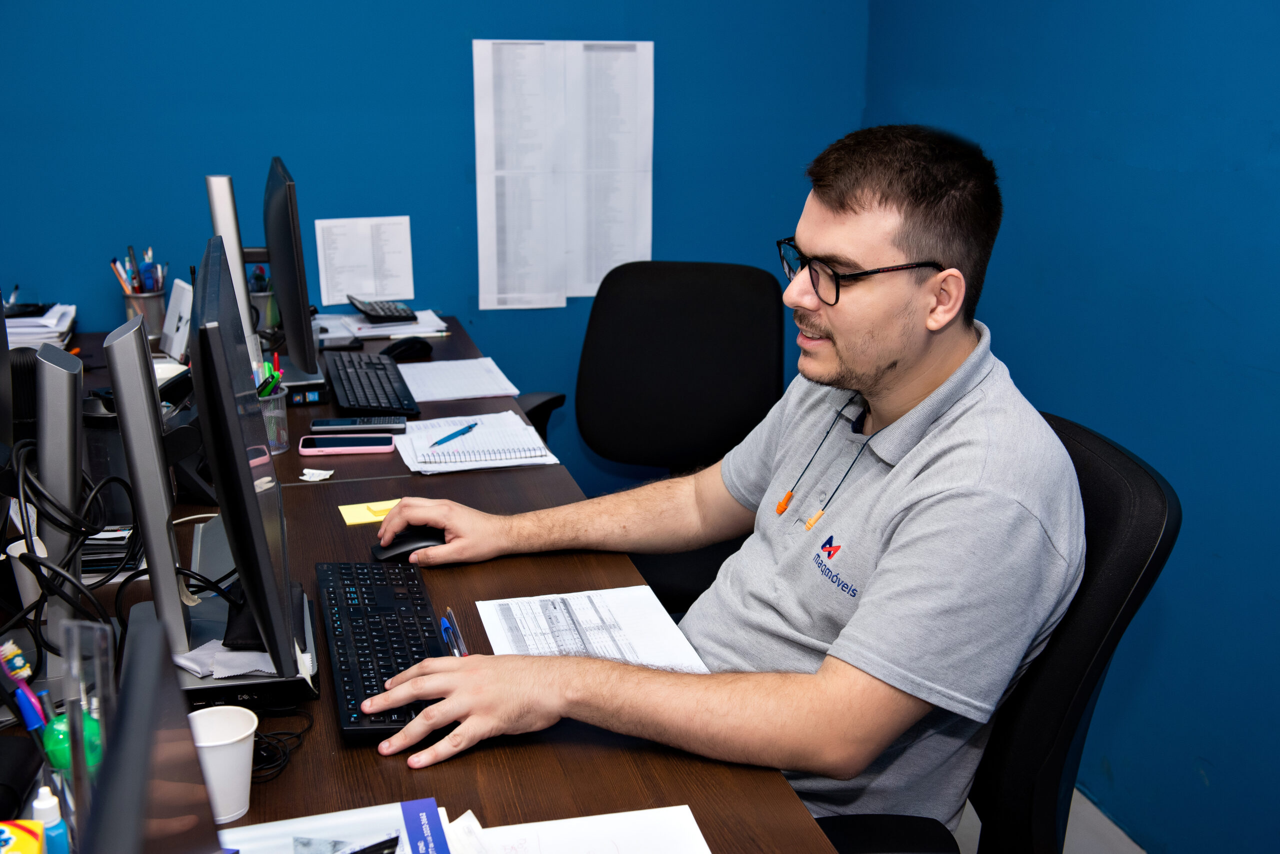 A imagem apresenta um homem branco, de cabelos escuros e curtos, que faz uso de óculos e uma camiseta cinza, em seu posto de trabalho, uma mesa com computador, em uma sala de paredes azuis.