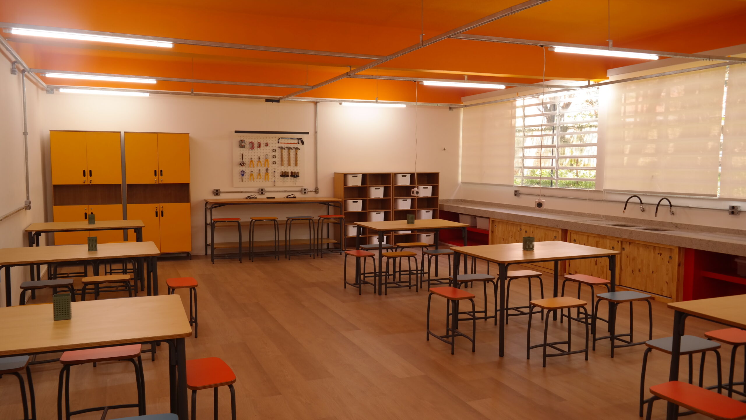 Sala de aula com mesas coletivas e bancos laminados de cores variadas. Ao fundo, armários e painel com ferramentas.