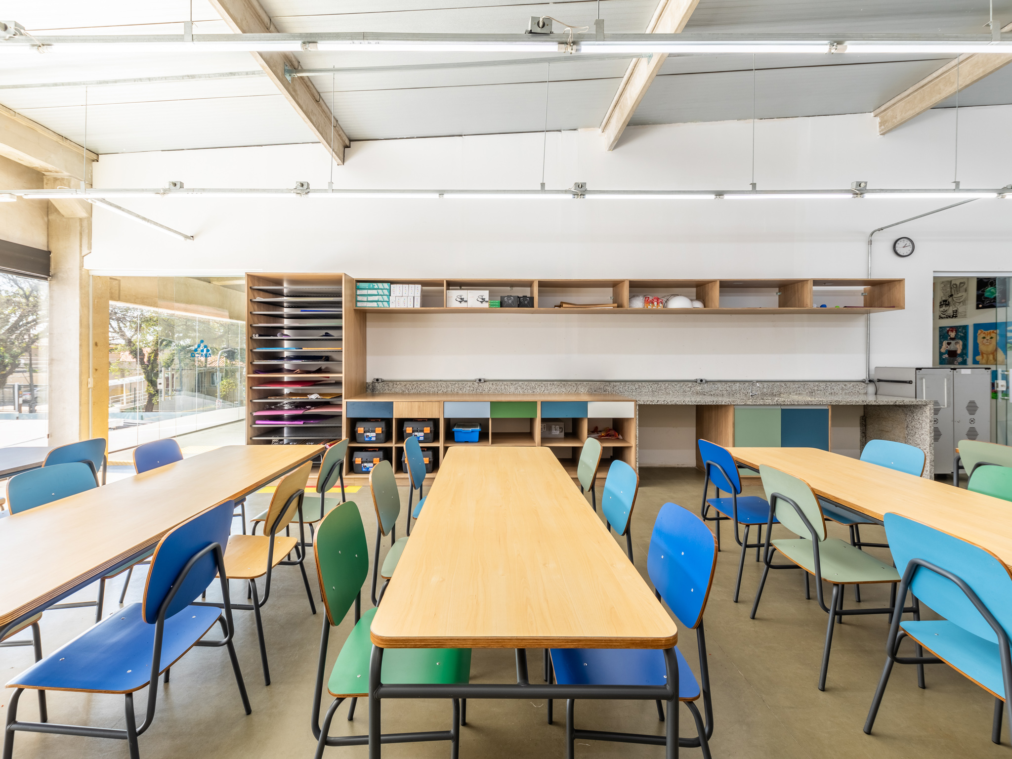 Sala de aula com mesas retangulares e cadeiras coloridas. No fundo, prateleiras com materiais escolares.
