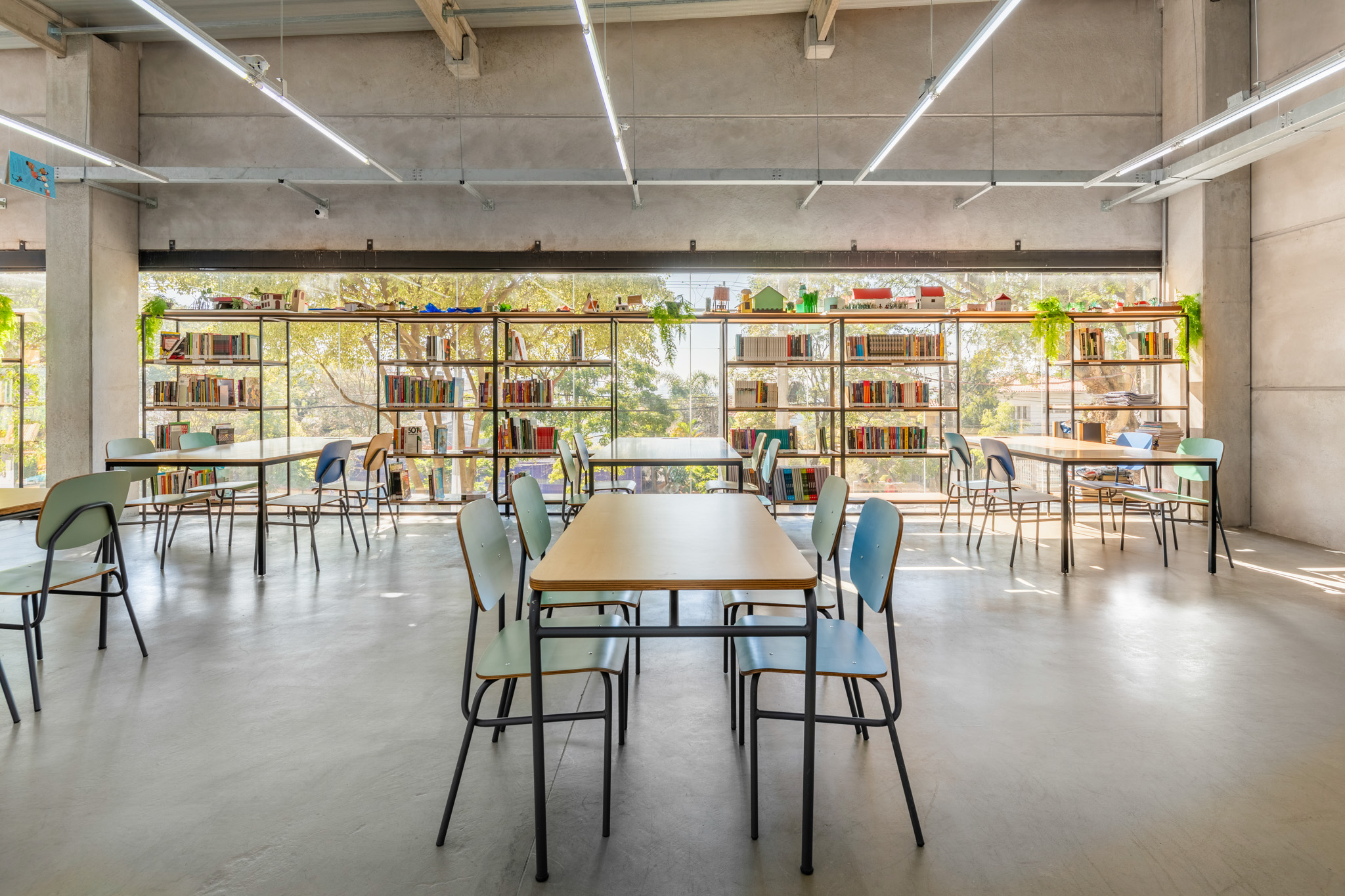 Sala de leitura ampla e bem iluminada, com grandes janelas, mesas retangulares, cadeiras coloridas e estantes de livros vazadas.