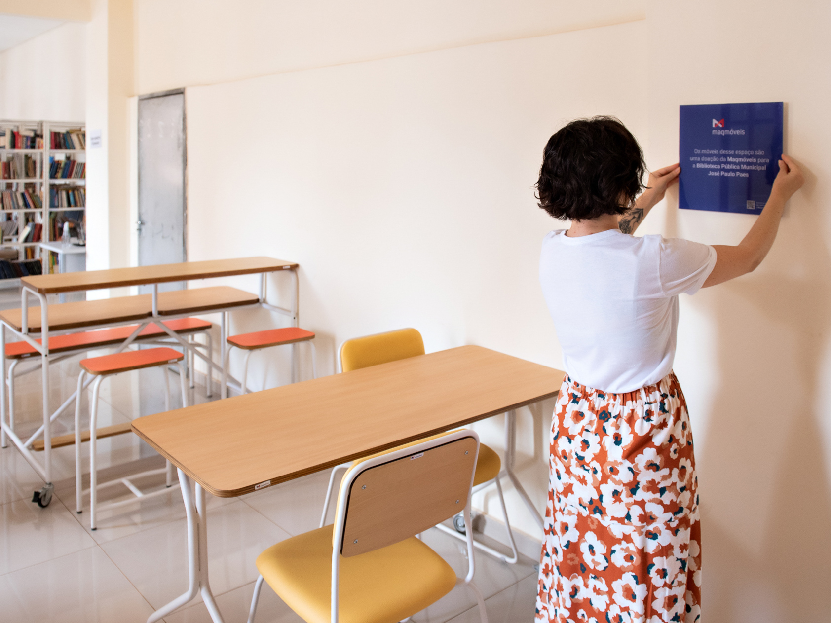 Sala de uma biblioteca com mesas e bancos coloridos. Uma mulher pendura um quadro na parede.