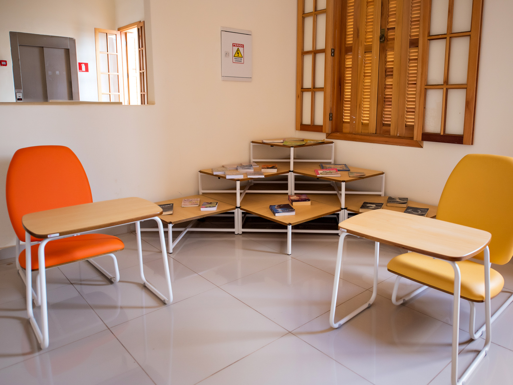 Sala de uma biblioteca com mesas e cadeiras coloridas. Alguns livros espalhados em uma das mesas.