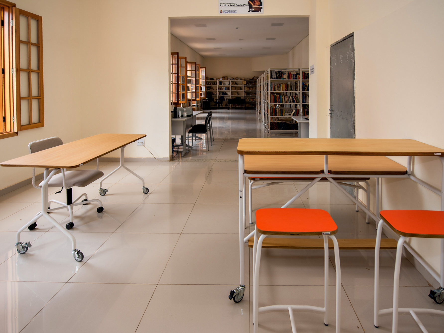 Sala de uma biblioteca com mesas e cadeiras coloridas. No fundo, várias estantes cheias de livros.