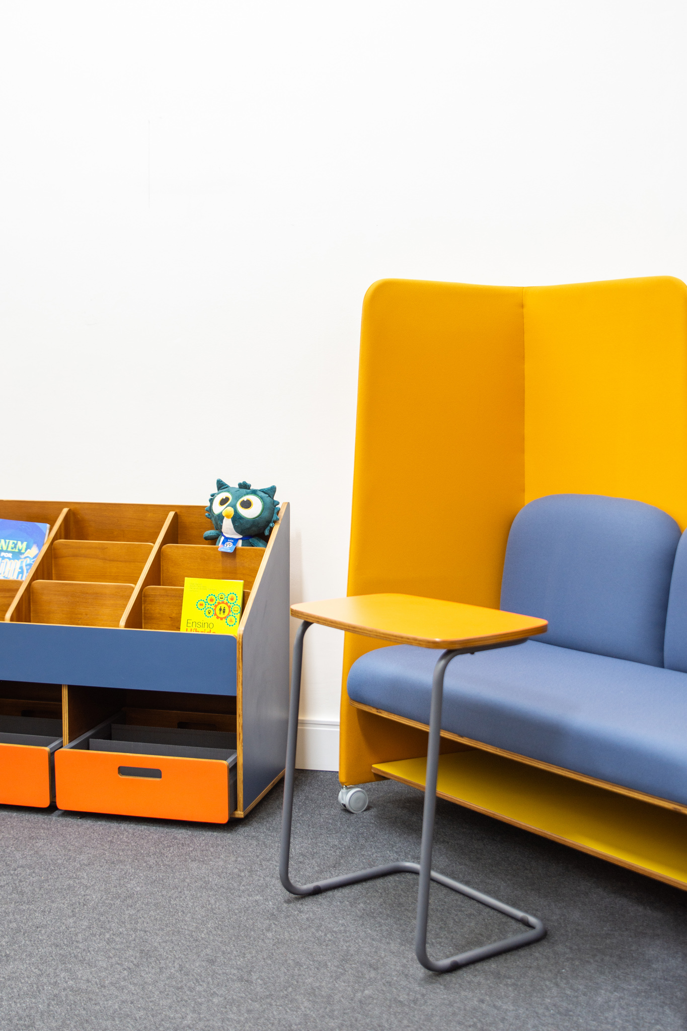 Sofá azul com painel de proteção amarelo, mesa de apoio lateral e uma estante de livros com gavetas laranjas, em um ambiente moderno.