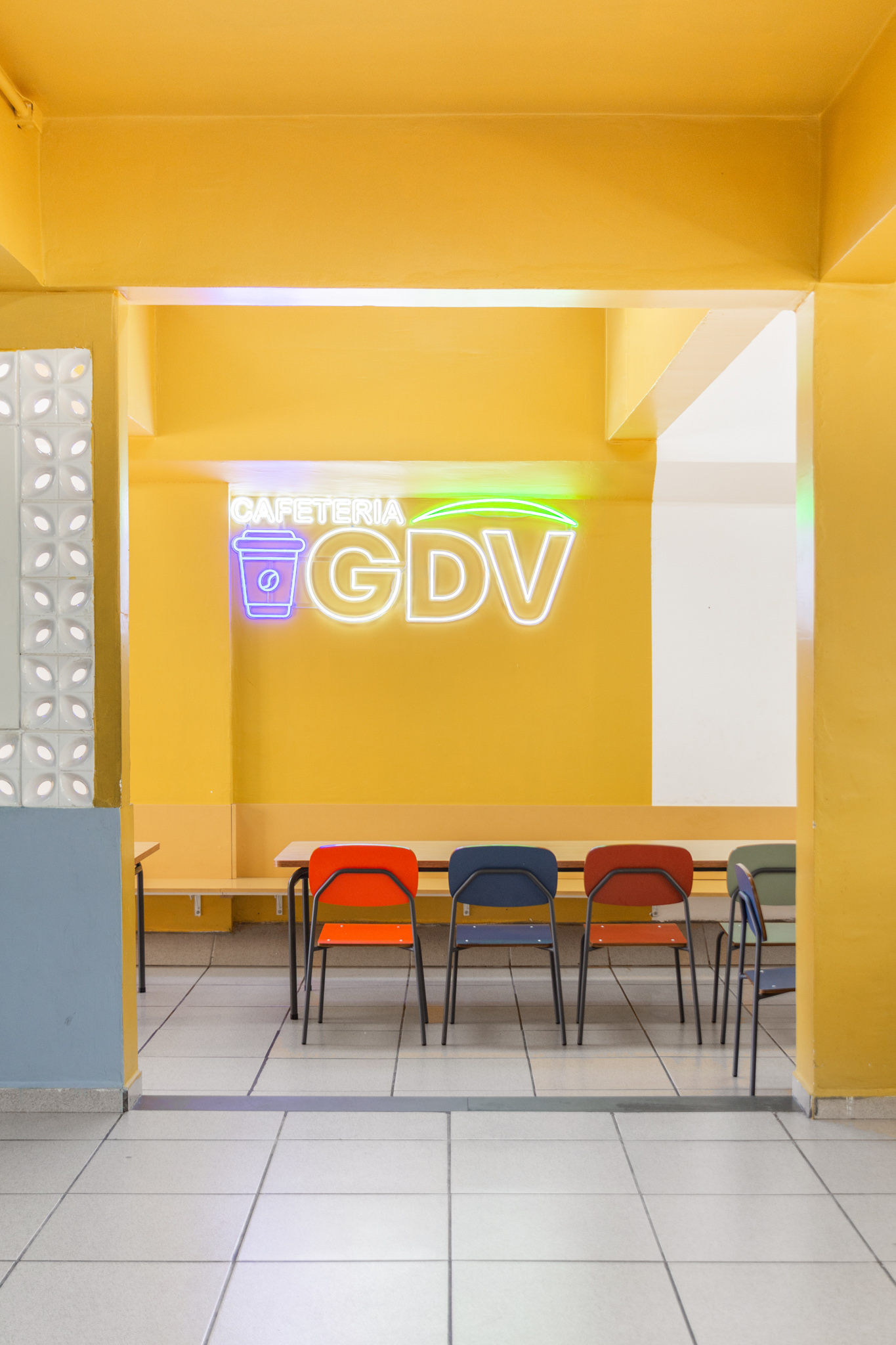 Cafeteria de escola com paredes amarelas e letreiro de neon, com cadeiras e bancos coloridos que representam móveis escolares modernos.