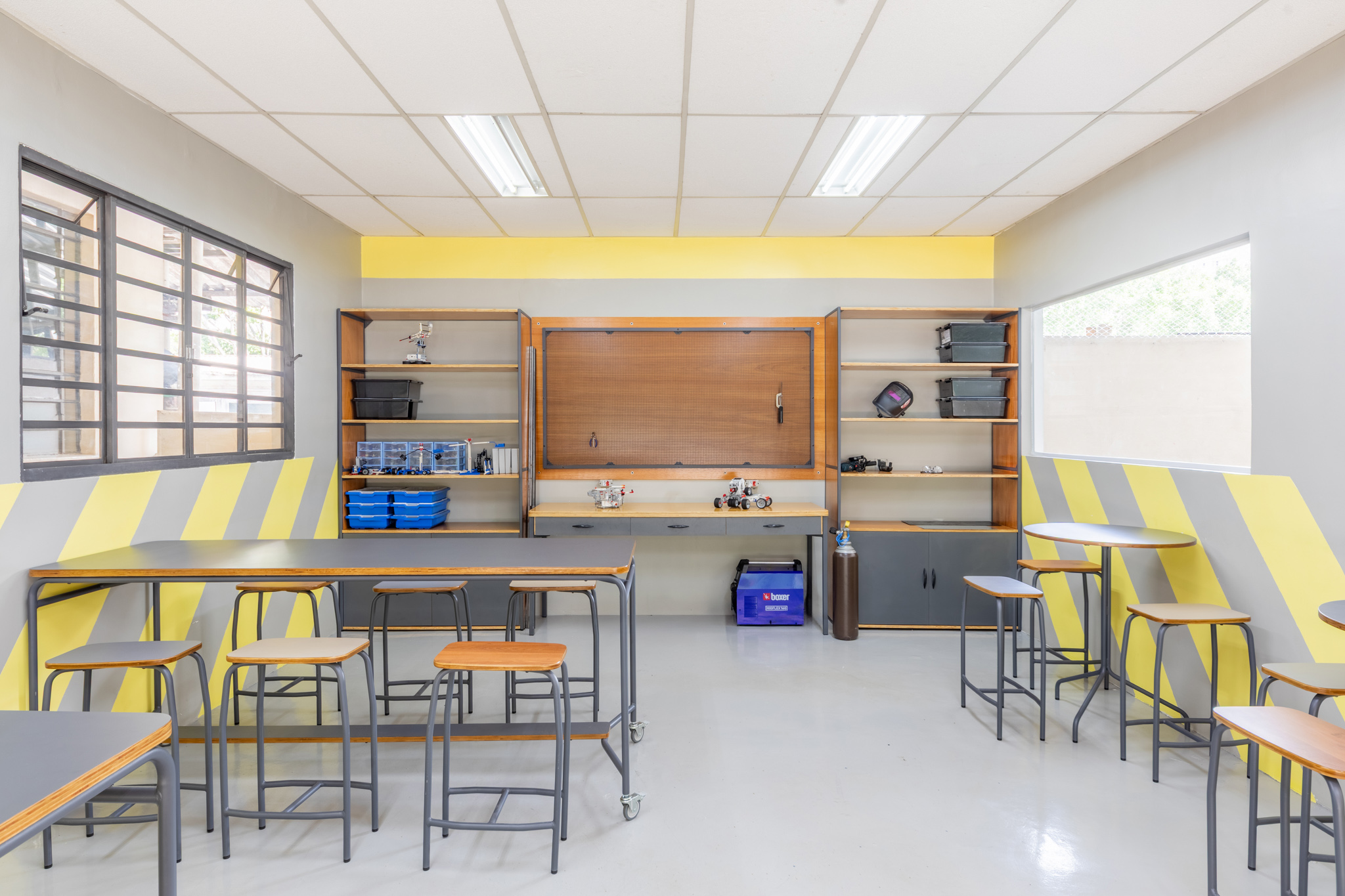 Sala de aula ou laboratório com mesas altas e baixas, bancos de design industrial e uma grande bancada de trabalho com prateleiras.