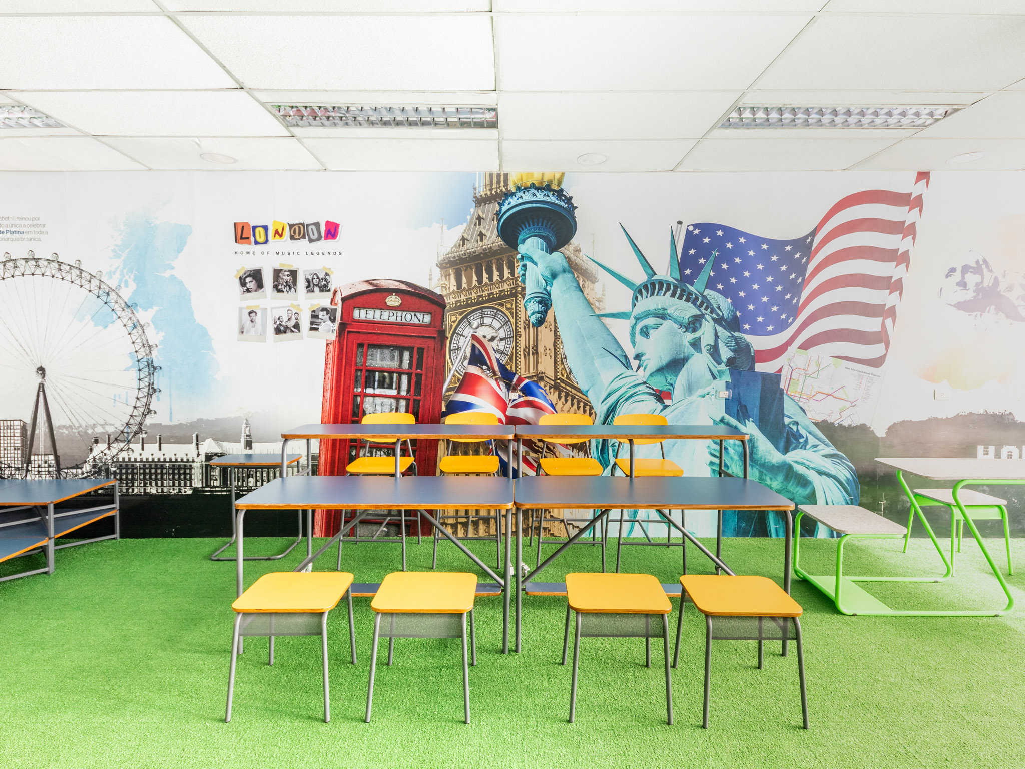 Ambiente de aprendizado com mesas e cadeiras modulares, sobre grama sintética, em sala com murais temáticos.