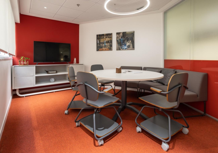 Sala de reuniões moderna e colorida com paredes brancas e vermelhas, poltronas confortáveis, mesas e uma televisão grande.