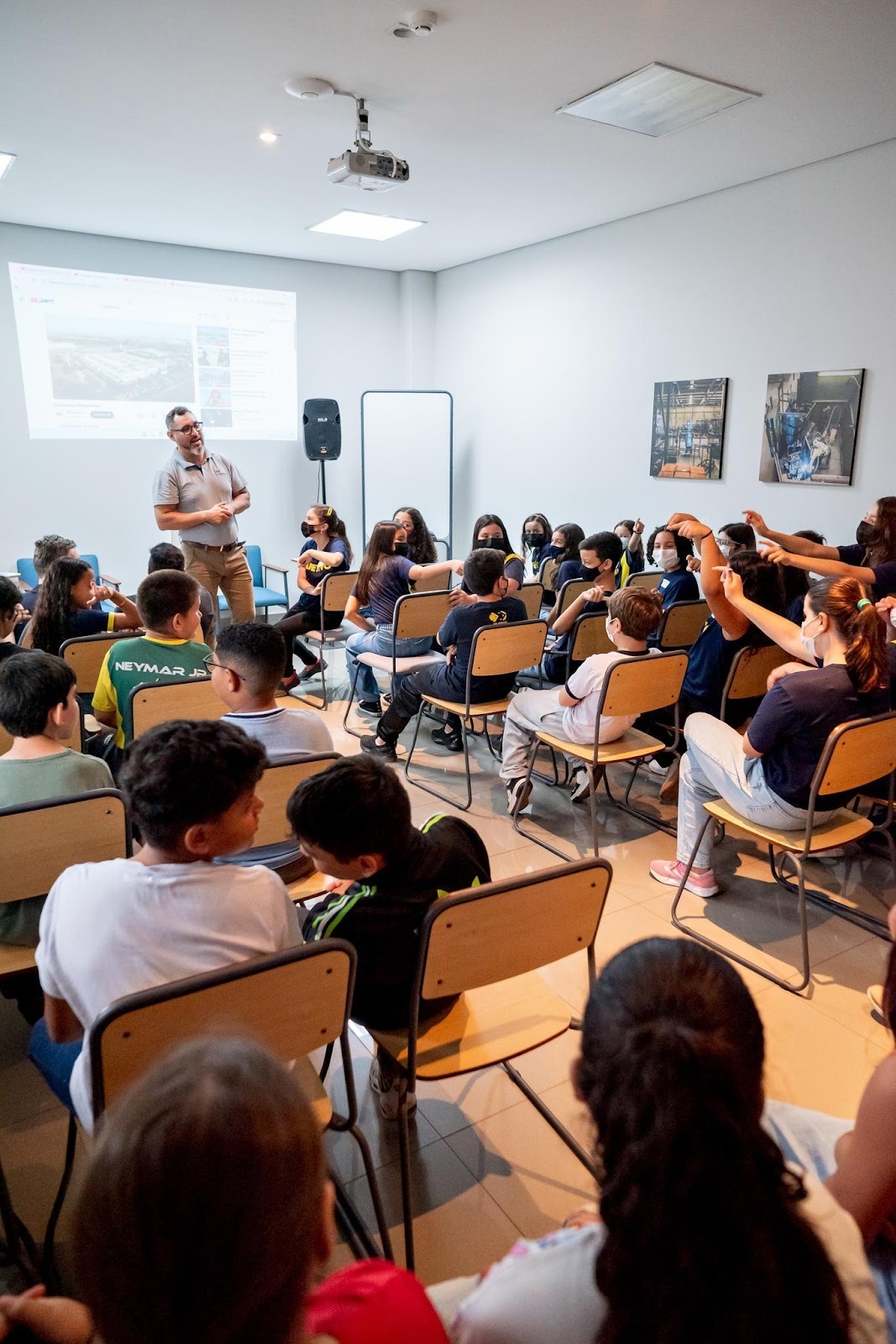 Professor em pé na frente de uma tela projetada, dando uma palestra para uma turma de jovens e adolescentes sentados em cadeiras em uma sala de aula.