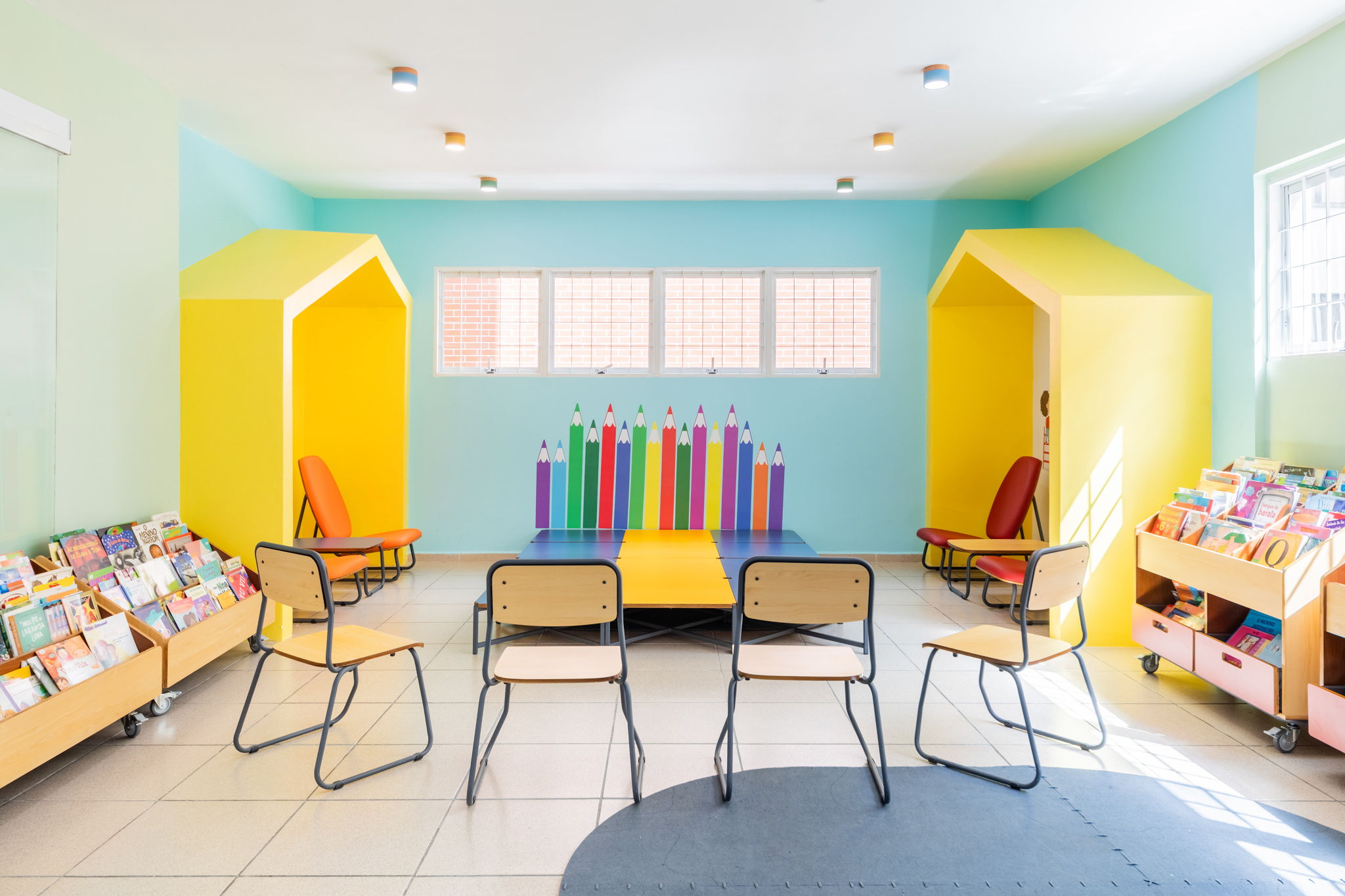 Sala lúdica com nichos amarelos em formato de casa, cadeiras de madeira e estantes baixas, apresentando móveis para biblioteca escolar coloridos.