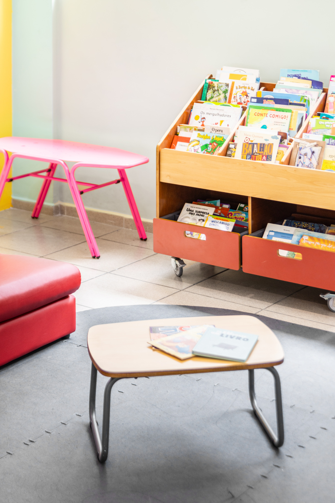 O Gemini disse Cantinho de leitura com estante de madeira repleta de livros, mesa pequena e banco rosa, destacando o uso de móveis para biblioteca escolar.